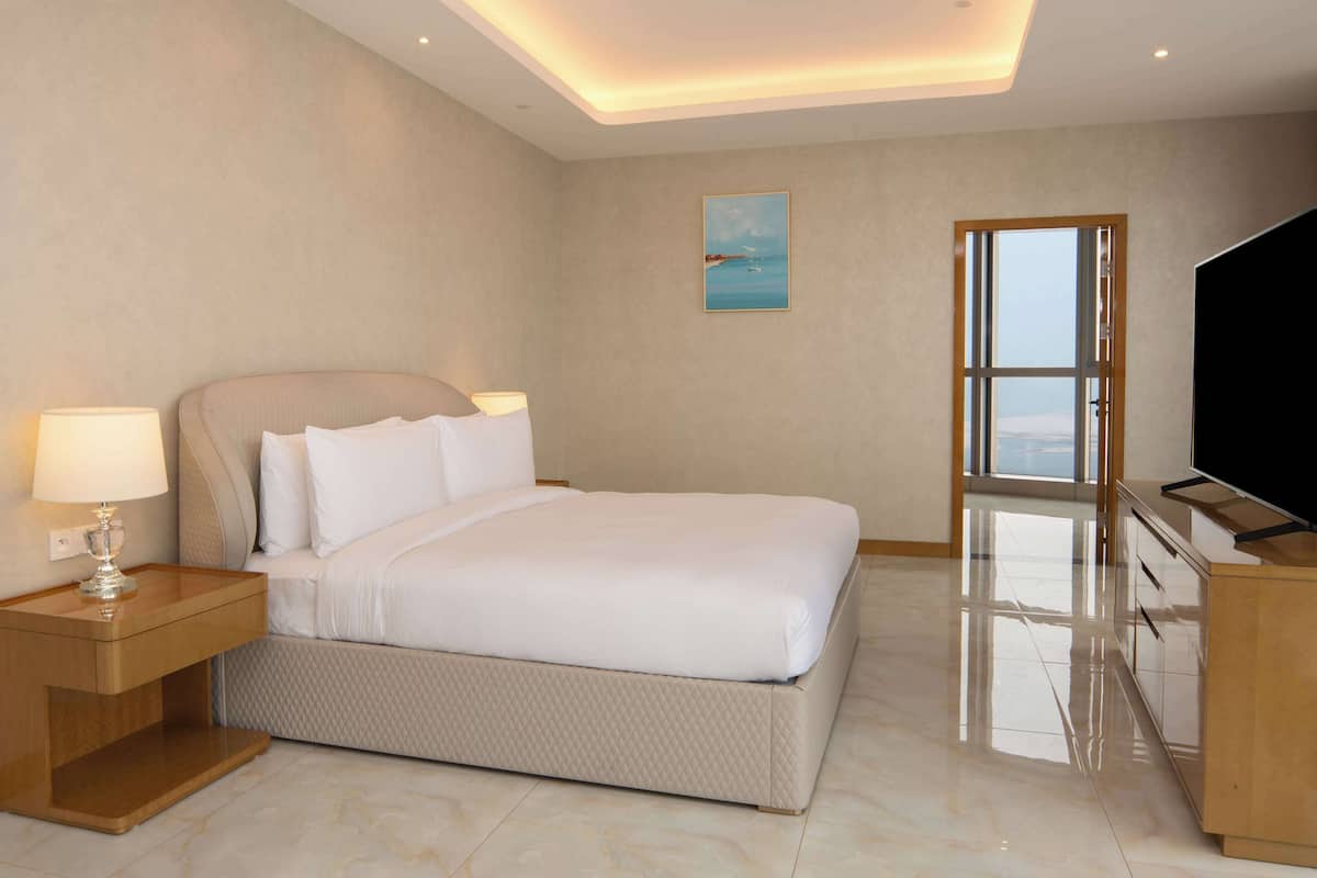 A modern hotel room with a large bed, a bedside table with a lamp, a flat-screen TV, and a large window offering a view of the sky.