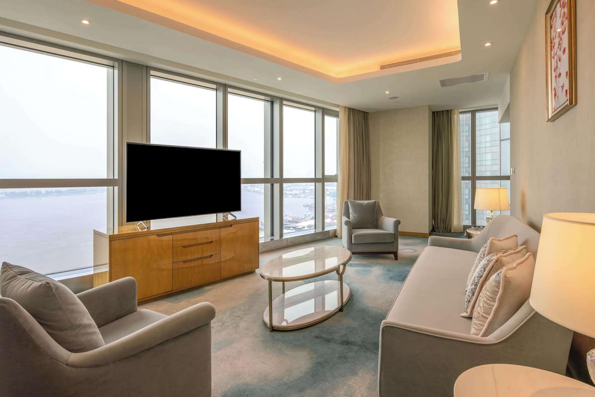 A modern hotel room with a large window, a flat-screen TV, a sofa, a chair, and a small table.