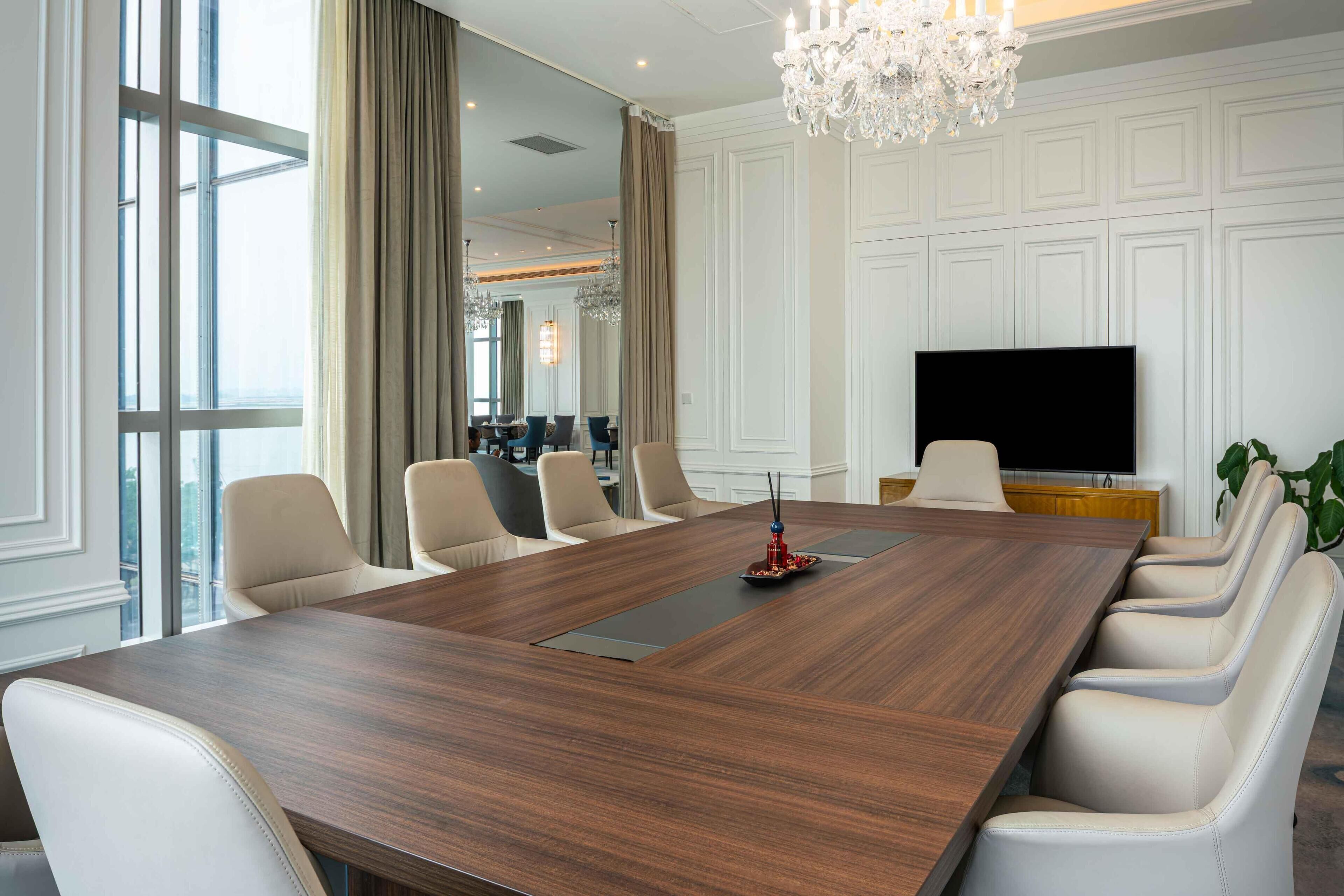 A conference room with a long wooden table, six chairs, a TV, and a chandelier.