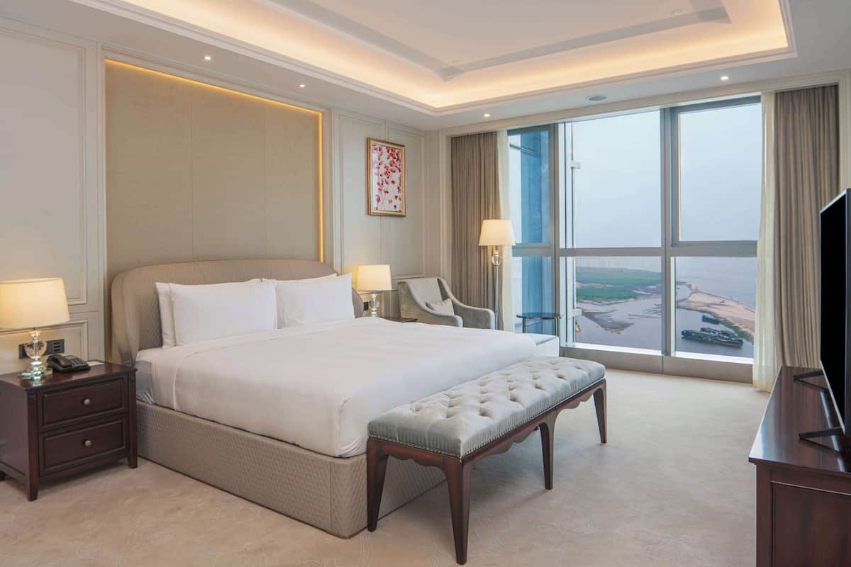 A hotel room with a large bed, a bedside table with a lamp, a chair, a television, and a large window with a view of a landscape.