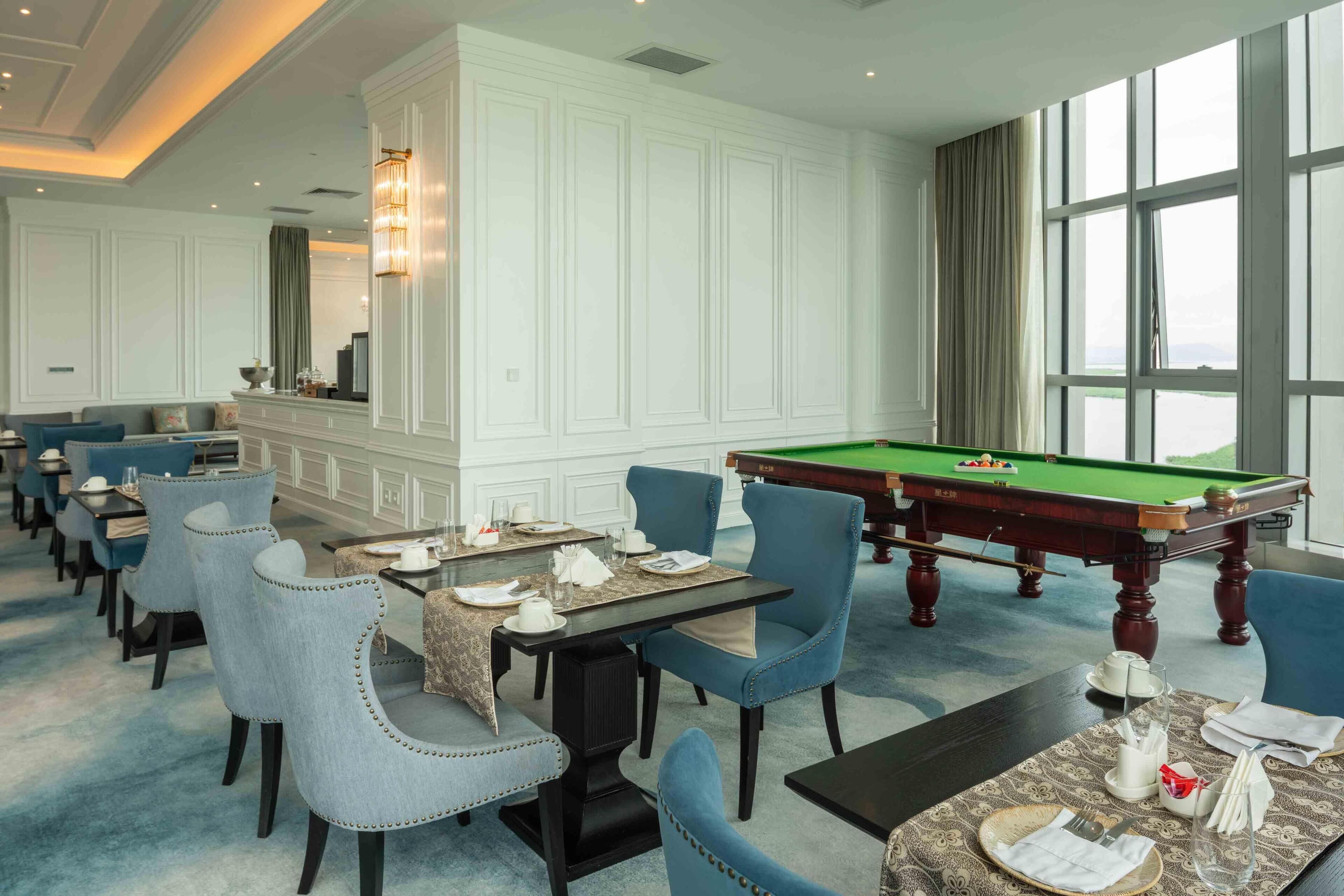 A hotel dining area with a pool table, multiple dining tables, and blue upholstered chairs.