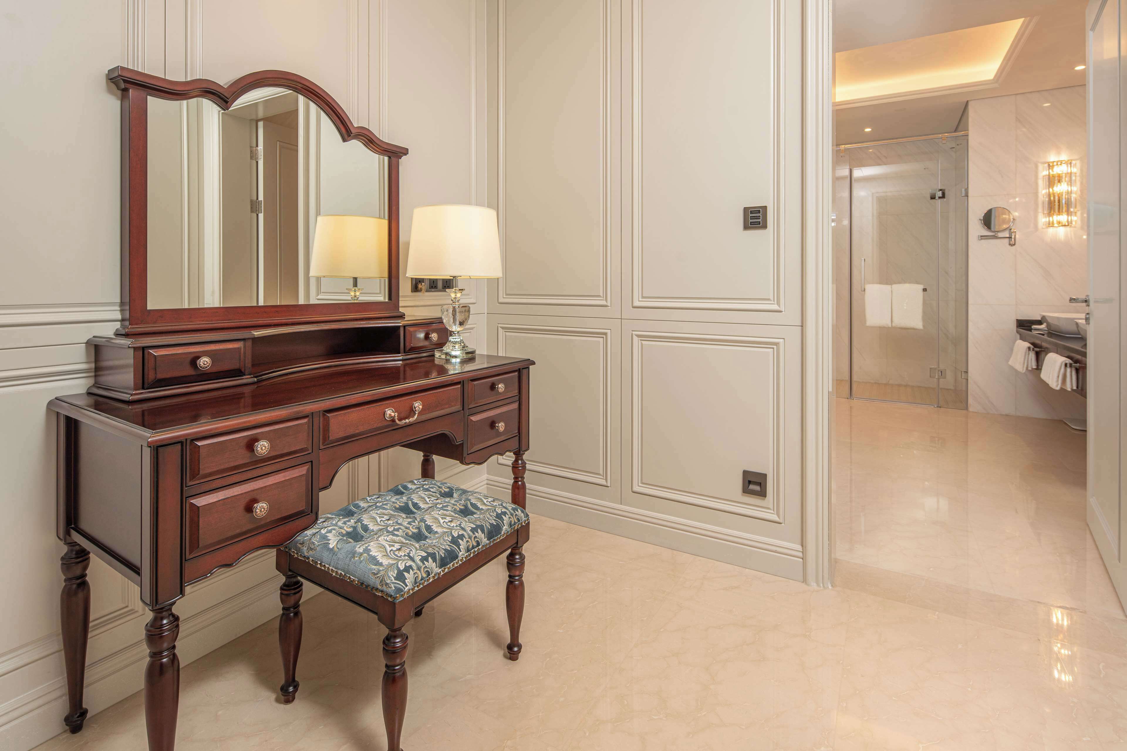 A wooden vanity with a mirror, two lamps, and a bench.