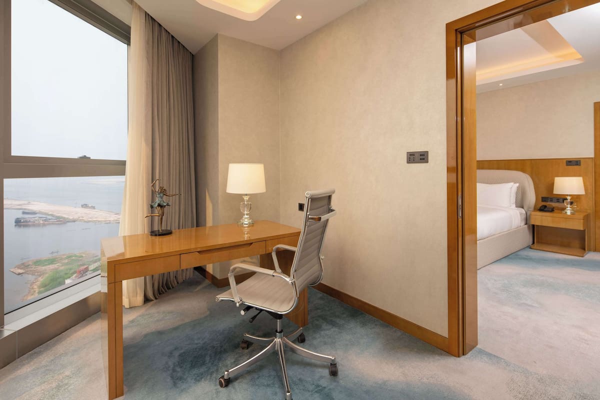 A hotel room with a desk, chair, bed, and a view of a body of water.