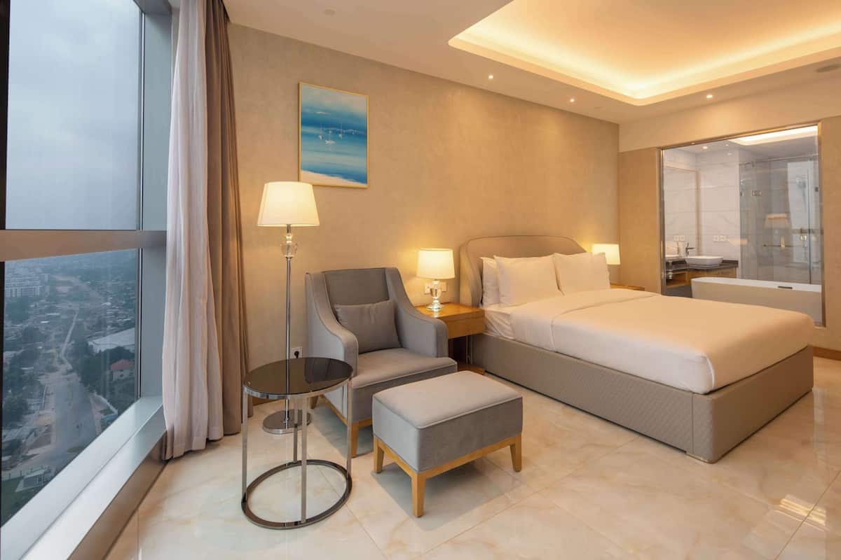 A modern hotel room with a large bed, a sitting area with a chair and table, a view of the city through the window, and a bathroom with a glass shower.