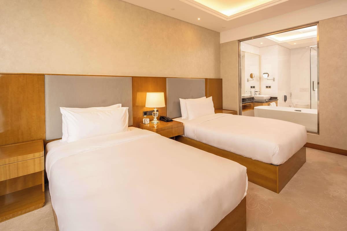 A hotel room with two single beds, a nightstand with a lamp, a mirror, and a bathroom visible through an open door.