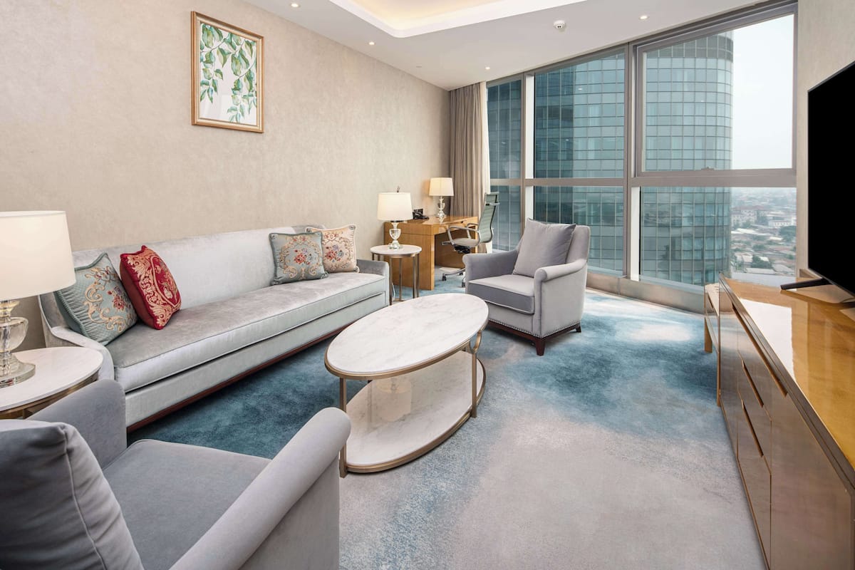 A modern hotel room with a grey sofa, a round coffee table, a grey armchair, a desk with a lamp, and a large window offering a city view.