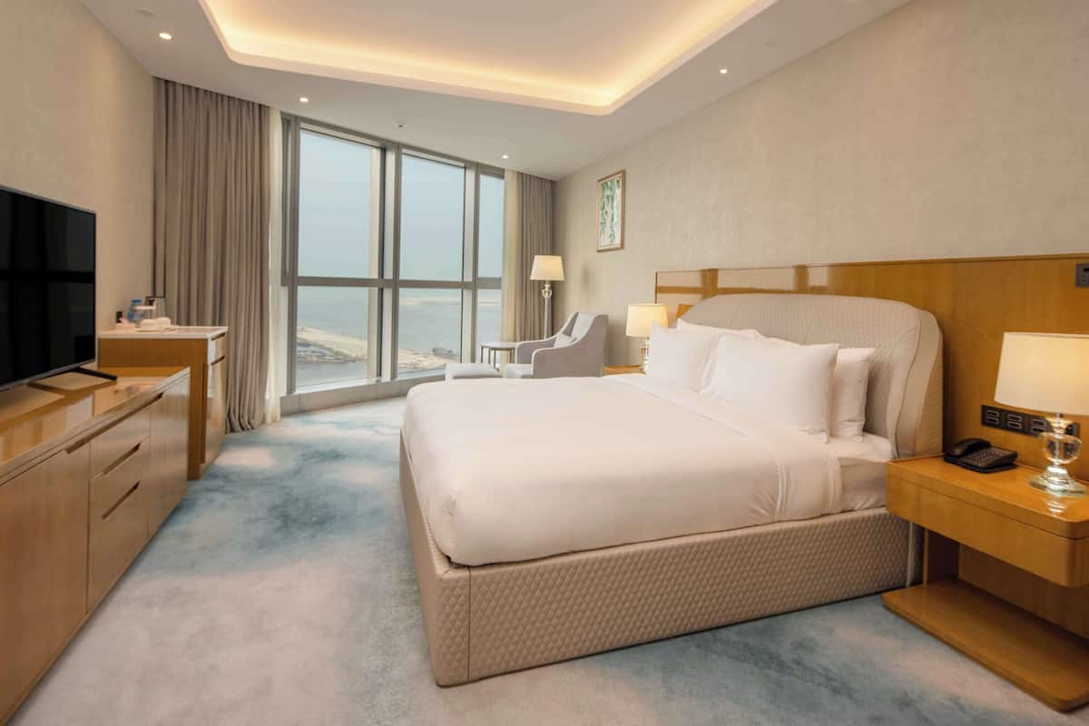 A hotel room with a large bed, a TV, a desk with a lamp, and a view of the sea.