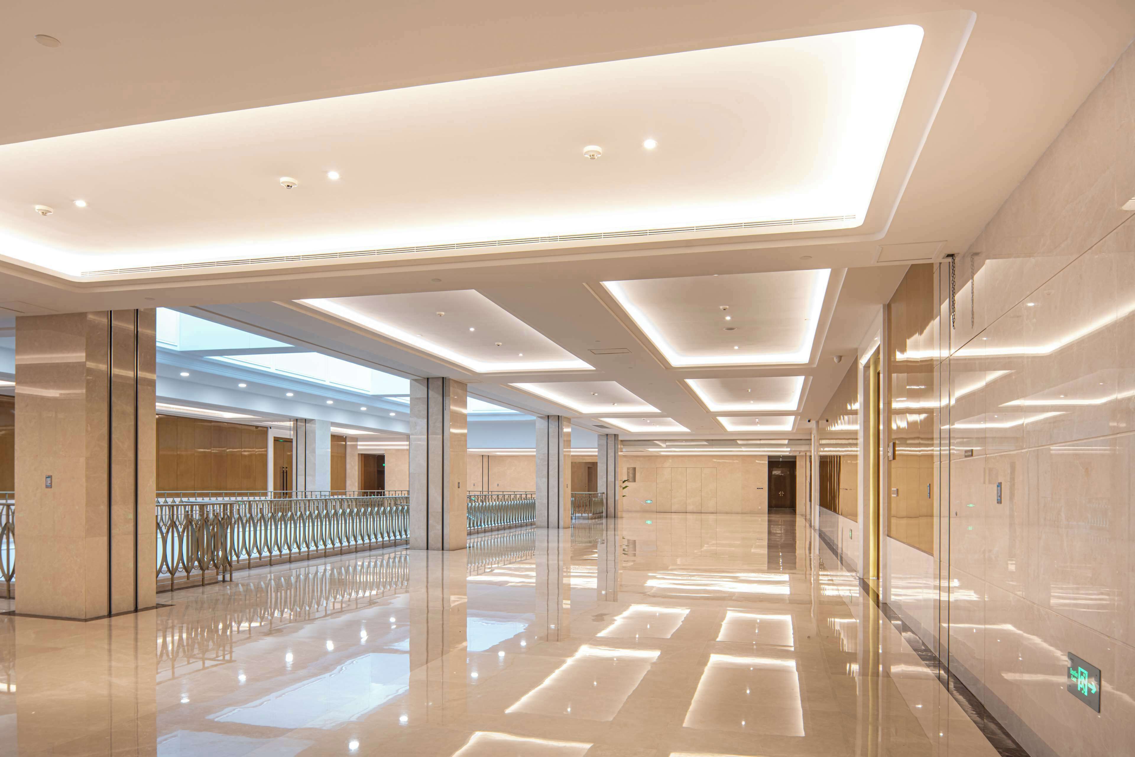 A spacious, modern lobby with a high ceiling, reflective flooring, and a glass railing overlooking a lower level.