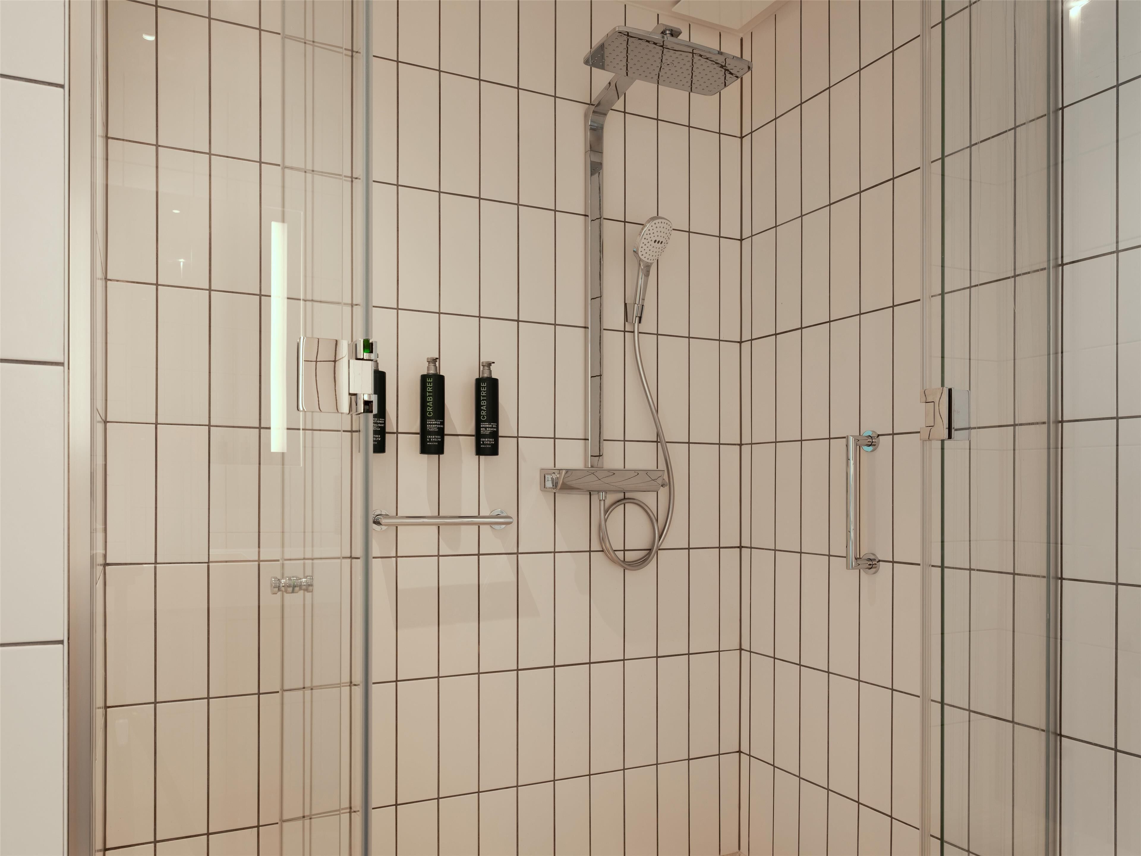 Un área de ducha moderna con puerta de vidrio, cabezal de ducha manual y dos botellas de gel de baño Crabtree.