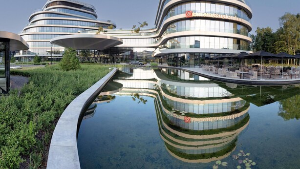 A modern hotel with a circular design, a reflection pool, and outdoor seating areas. - Hilton Garden Inn Brussels Airport (Machelen)