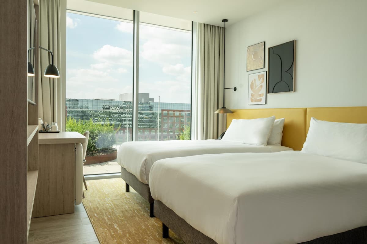 Ein Hotelzimmer mit zwei Betten, einem Schreibtisch und einem großen Fenster mit Blick auf die Stadt.