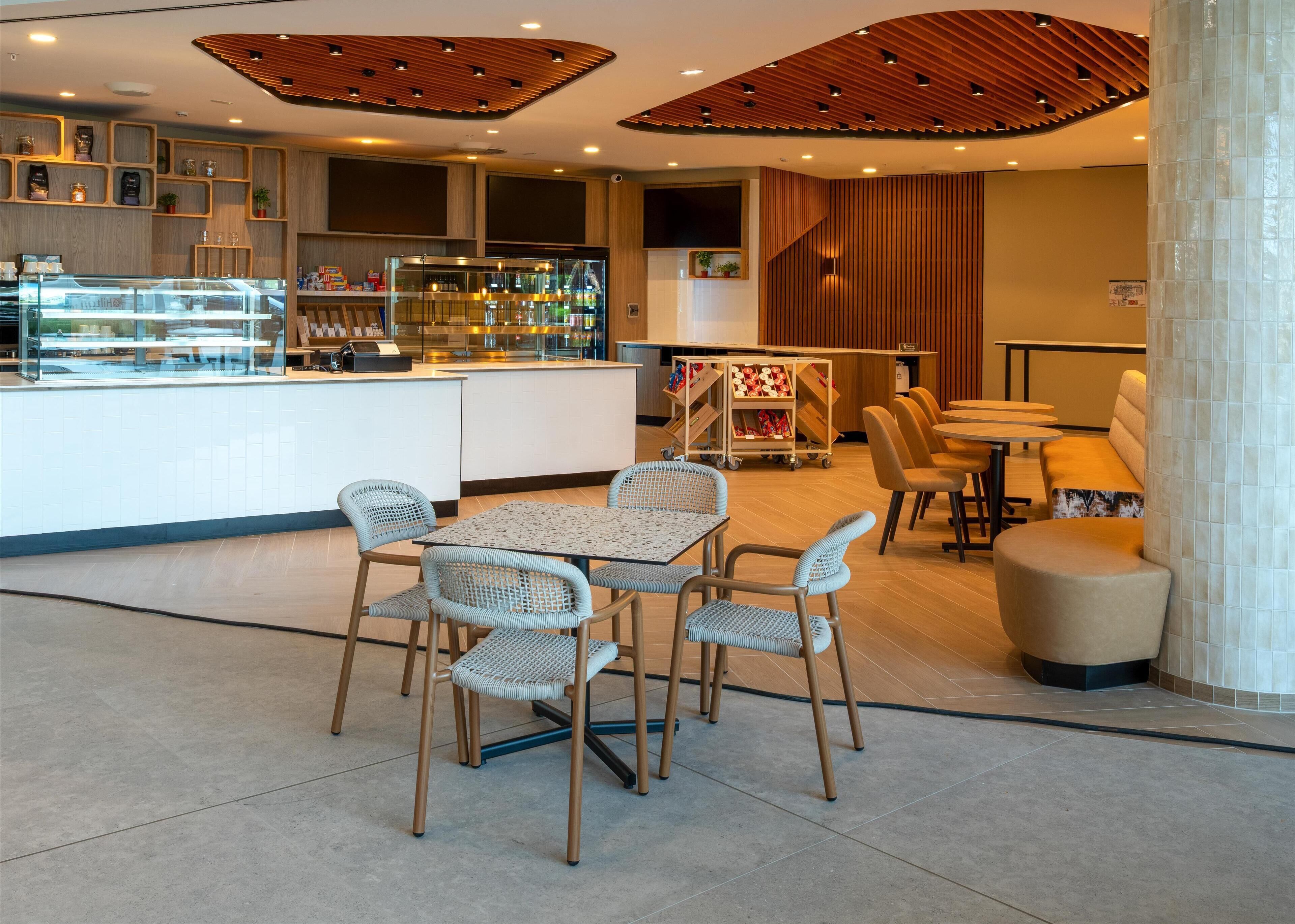 A modern cafe with a central island counter, wicker chairs, and a seating area with tables and sofas.