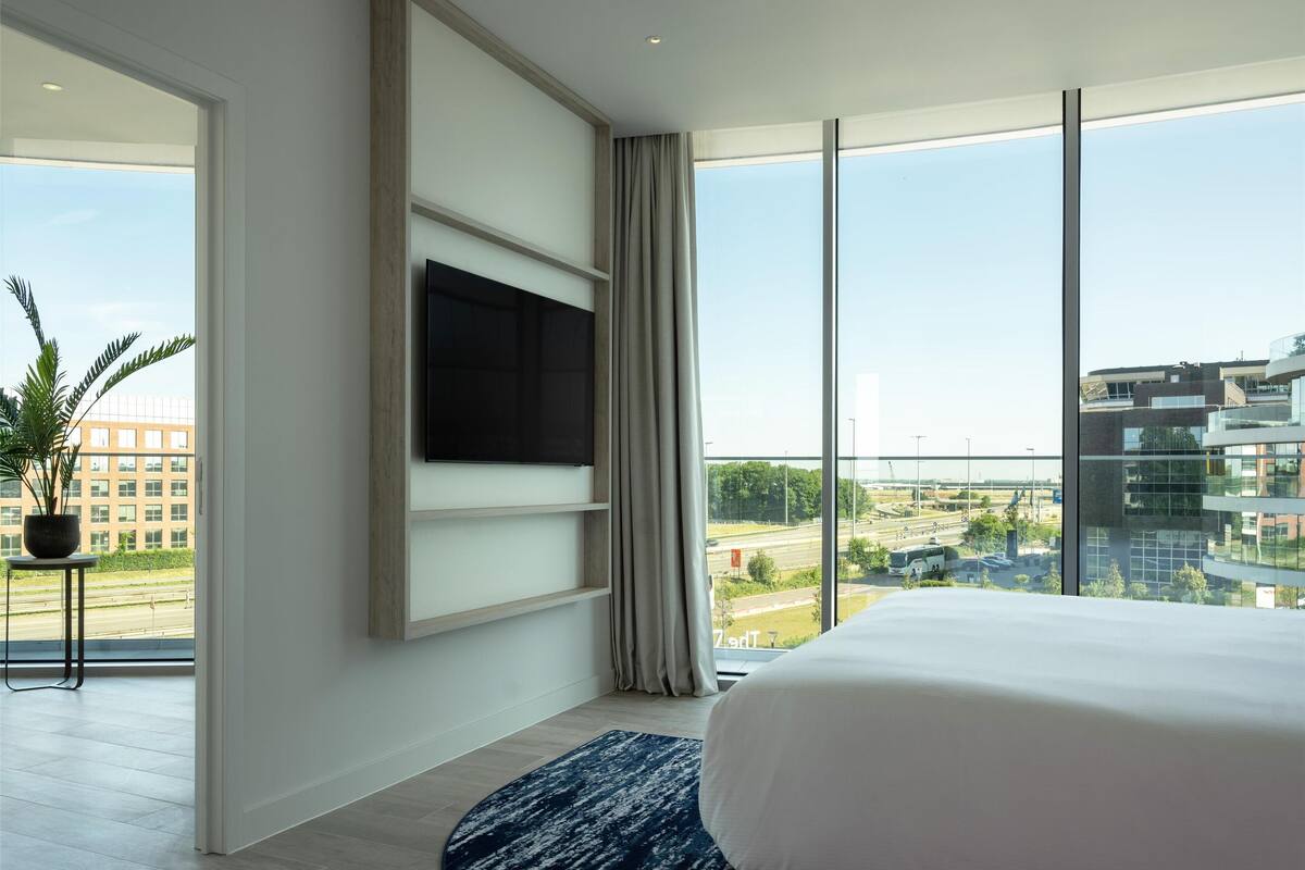 Ein modernes Hotelzimmer mit einem großen Bett, einem flachbildfernseher an der Wand, einer Topfpflanze und Blick auf die Stadt durch große Fenster.