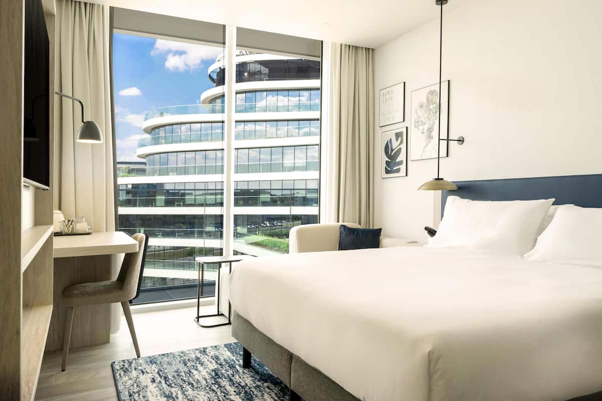 Ein modernes Hotelzimmer mit einem großen Bett, einem Schreibtisch und einem Sessel. Ein großes Fenster bietet Blick auf ein Hochhaus.