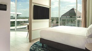 A modern hotel room with a large bed, a flat-screen TV mounted on the wall, and a view of the cityscape through large windows. - Hilton Garden Inn Brussels Airport (Machelen)