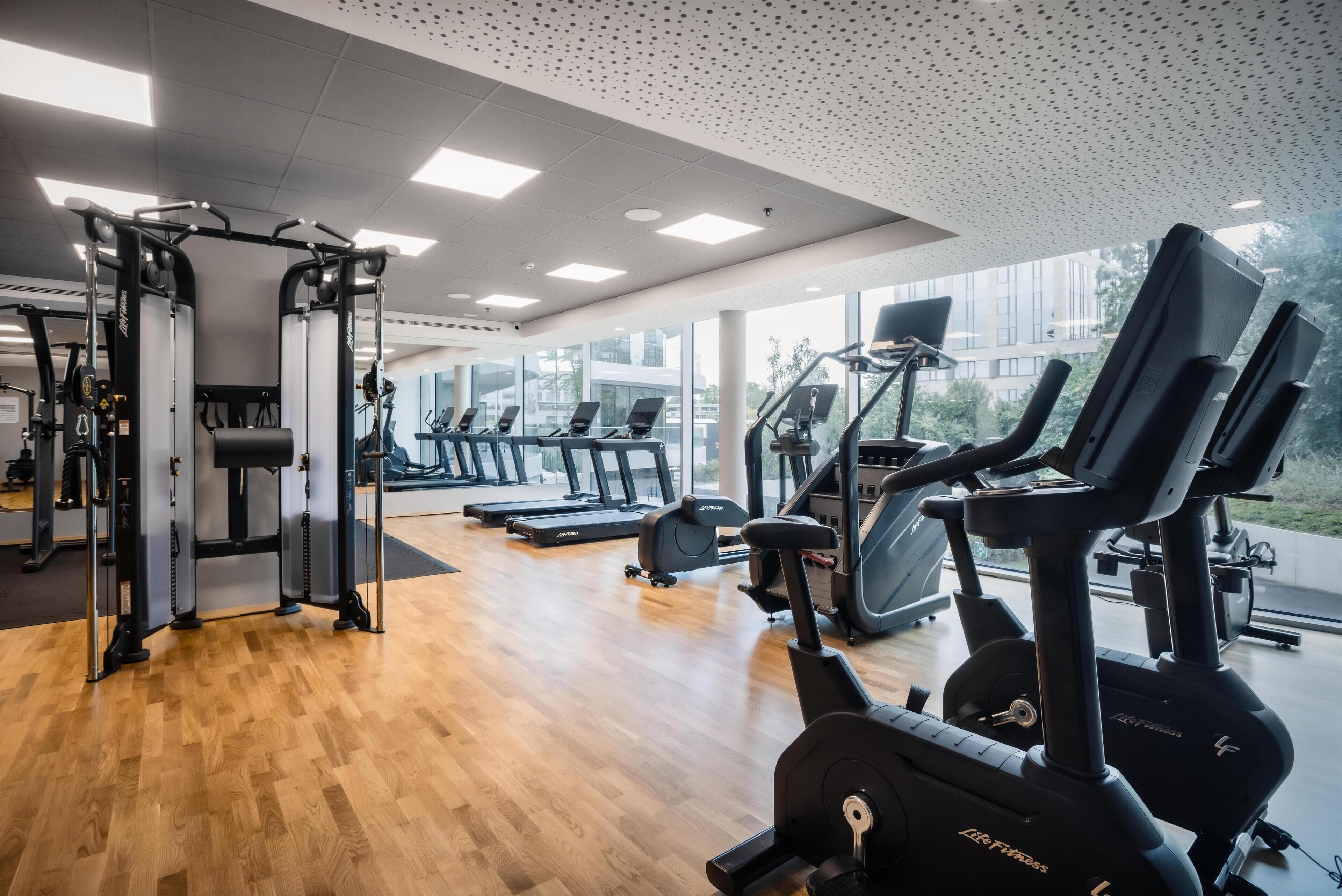 Une salle de sport moderne équipée de machines de cardio, d’appareils de musculation et de grandes fenêtres offrant une vue sur la ville.