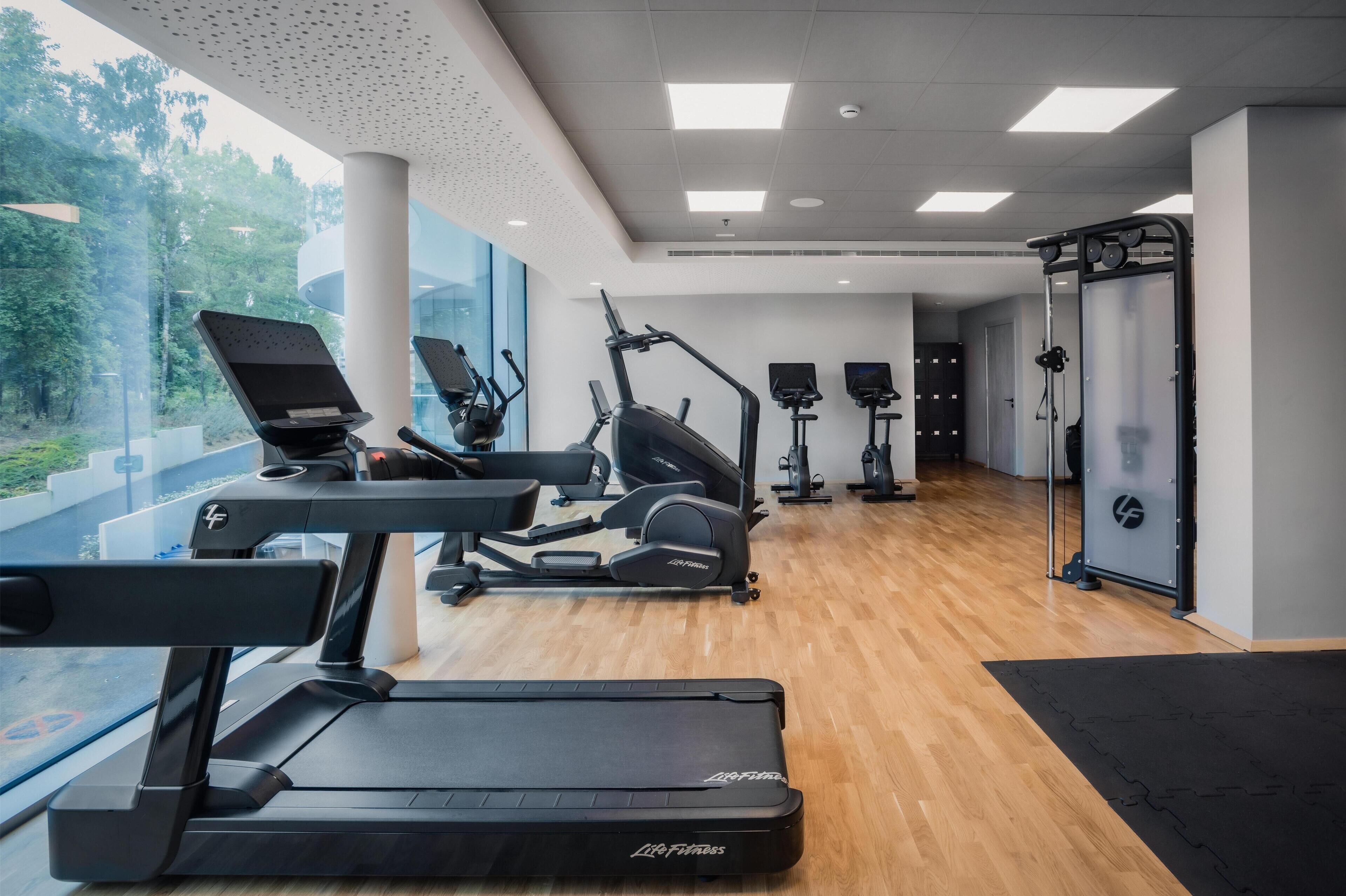 A modern gym with treadmills, elliptical machines, and exercise bikes. Large windows offer views of greenery outside.