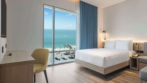 A hotel room with a bed, a desk, a chair, a sofa, and a large window with a view of the beach and ocean. - Hampton By Hilton Kuwait Salmiya (Salmiya)