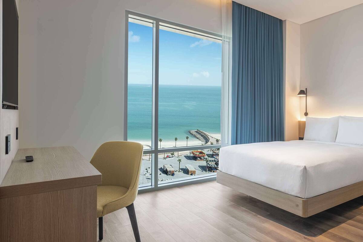 A hotel room with a bed, a desk, a chair, a sofa, and a large window with a view of the beach and ocean.