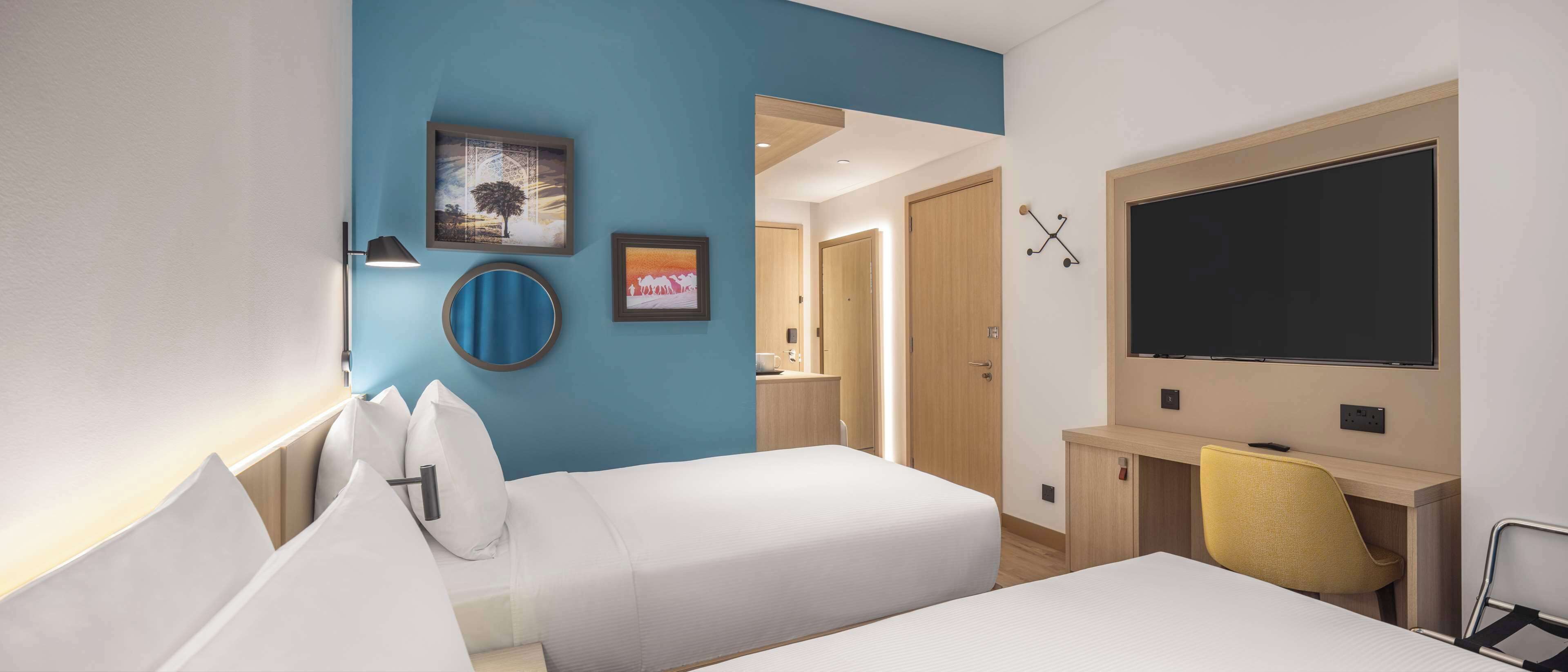 A hotel room with two beds, a desk, a chair, a TV, and artwork on the walls.