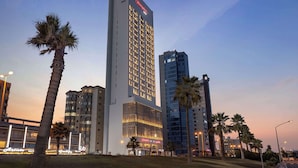 A tall hotel with a prominent 'Hampton' sign, surrounded by palm trees and other buildings. - Hampton By Hilton Kuwait Salmiya (Salmiya)