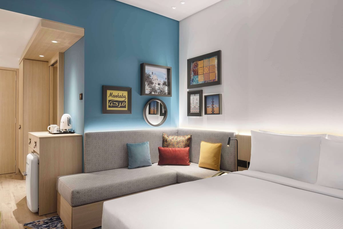 A modern hotel room with a grey sofa, a bed, a small desk with a kettle, and wall art.