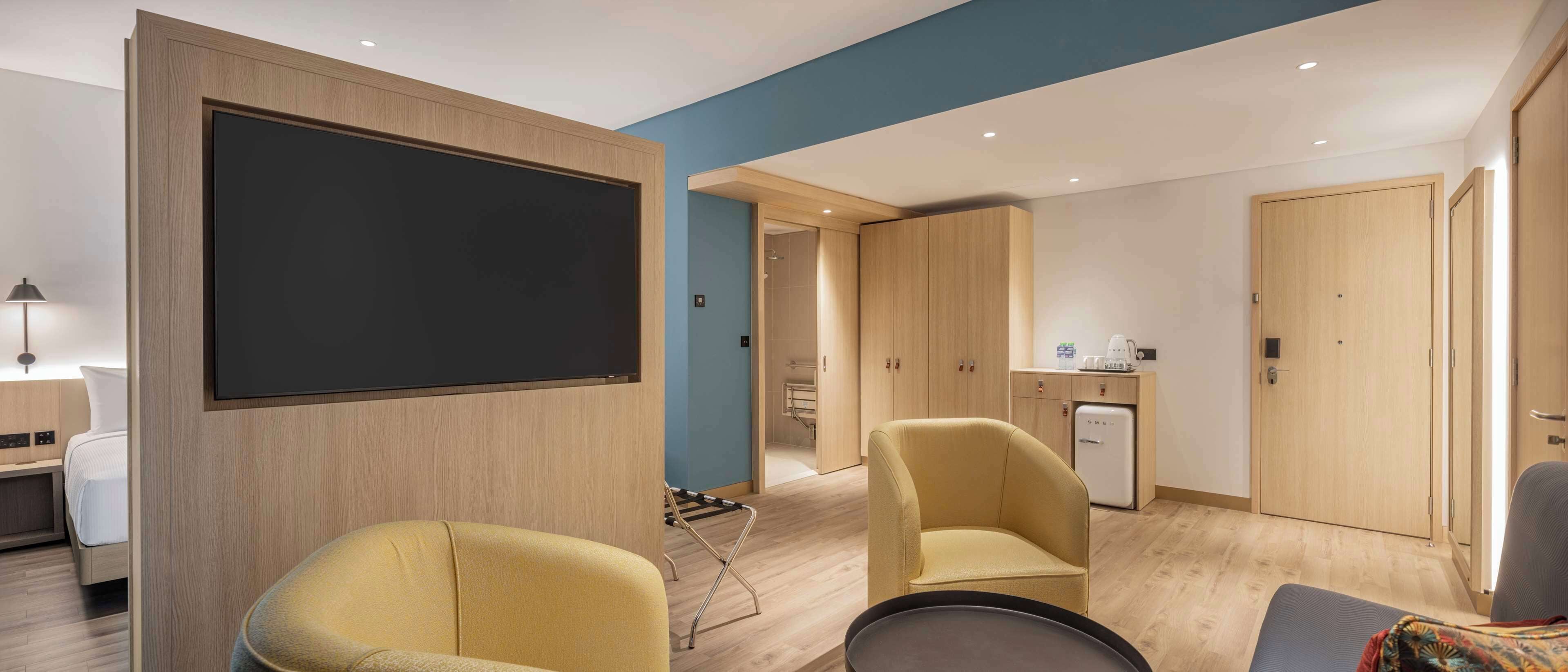 A modern hotel room with a large flat-screen TV, two yellow armchairs, a small table, and a compact kitchenette.