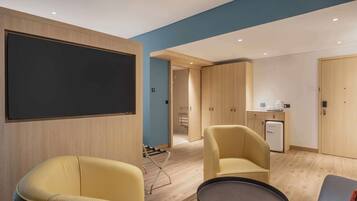 Una habitación de hotel moderna con un televisor de pantalla plana grande, dos sillones amarillos, una mesita pequeña y una kitchenette compacta.
