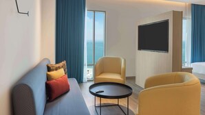 A modern hotel room with a sofa, two armchairs, a small table, a flat-screen TV, and a large window with a view of the sea. - Hampton By Hilton Kuwait Salmiya (Salmiya)