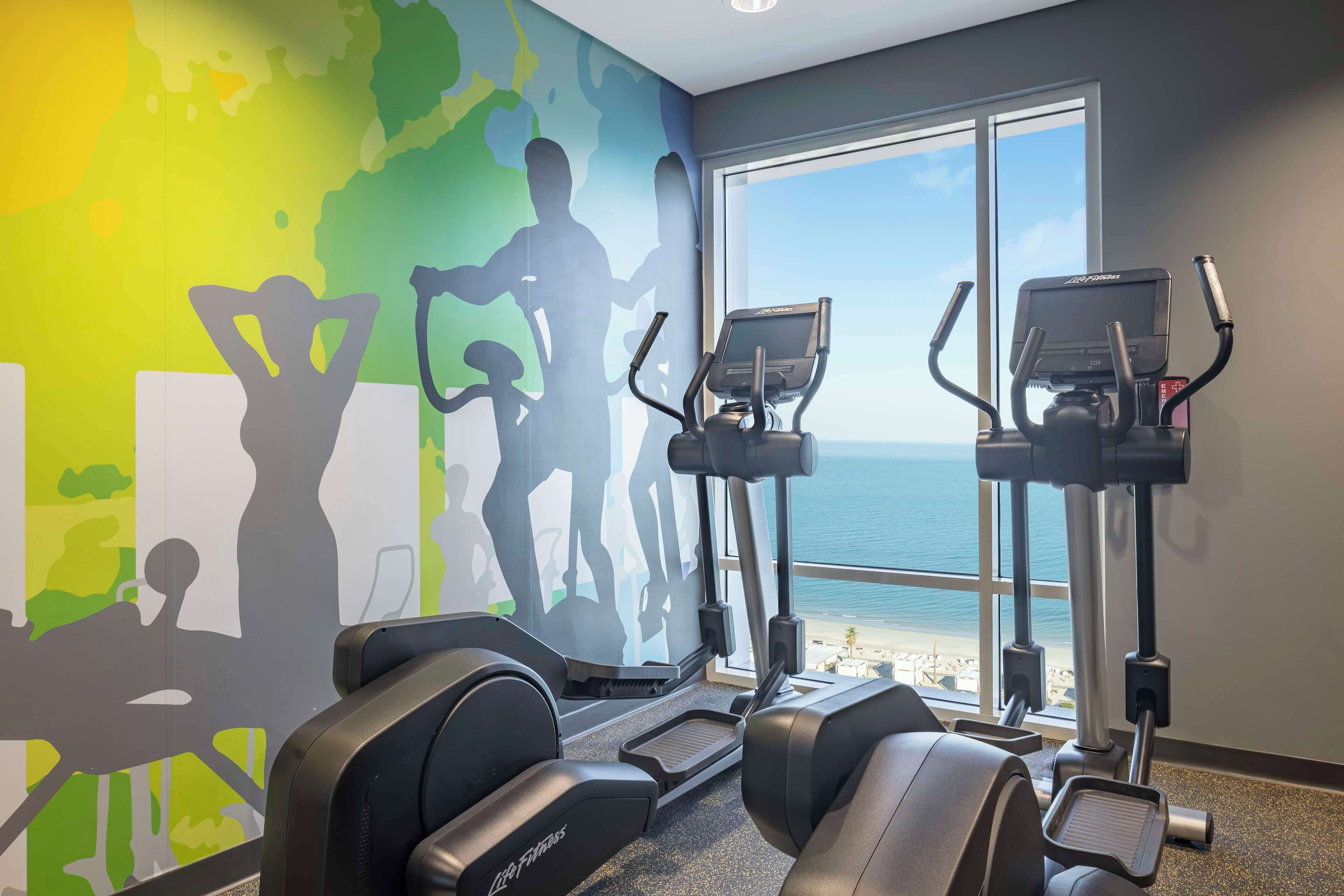 A gym with treadmills and elliptical machines, large windows with a view of the beach, and a colorful wall mural.