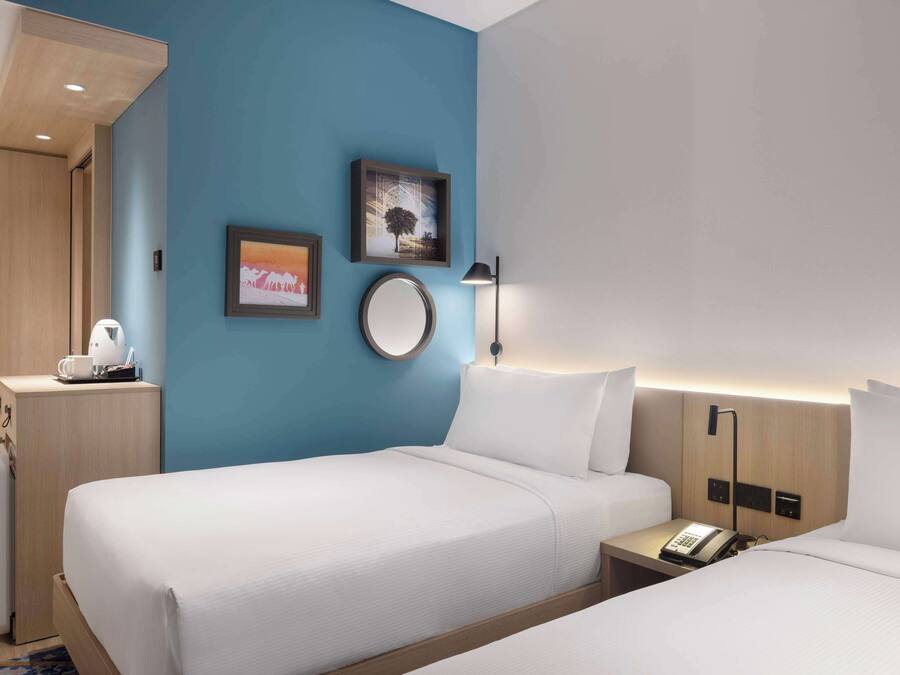 A hotel room with two beds, a desk with a lamp, and a small kitchenette.