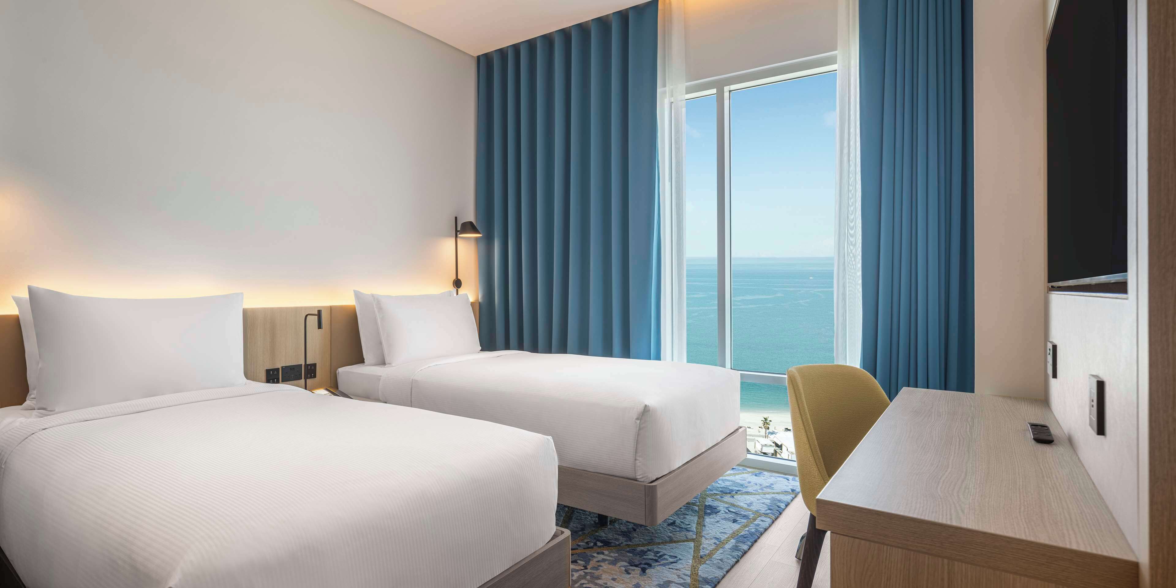 Habitación de hotel con dos camas, un escritorio, una silla y un ventanal con vista al mar.