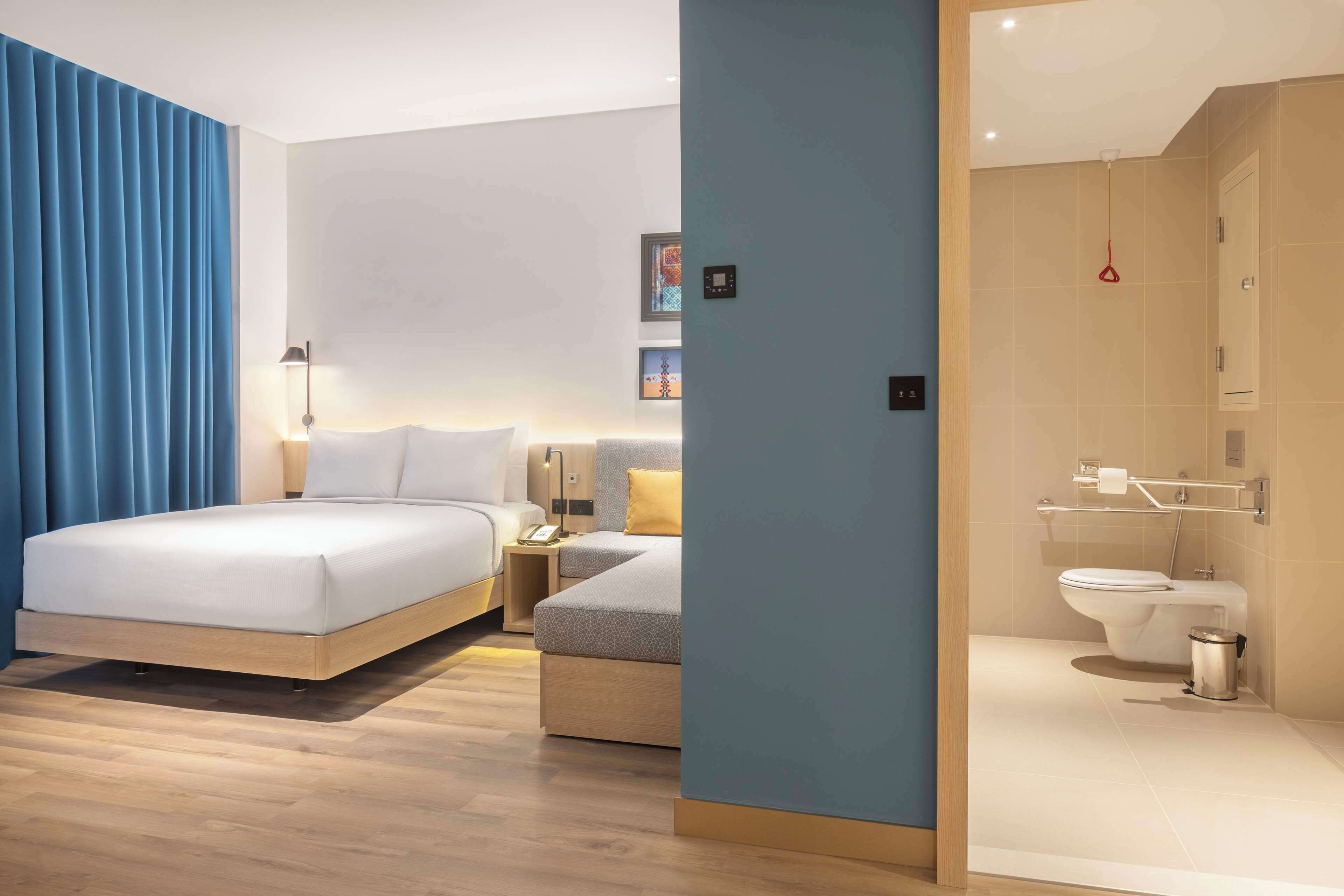 A modern hotel room with a large bed, a smaller sofa, a bedside table, a nightstand, a lamp, a wall-mounted TV, and a bathroom with a toilet and sink.