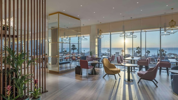 A modern indoor space with large windows offering a view of the sea, furnished with tables and chairs, and featuring contemporary lighting fixtures. - Hampton By Hilton Kuwait Salmiya (Salmiya)