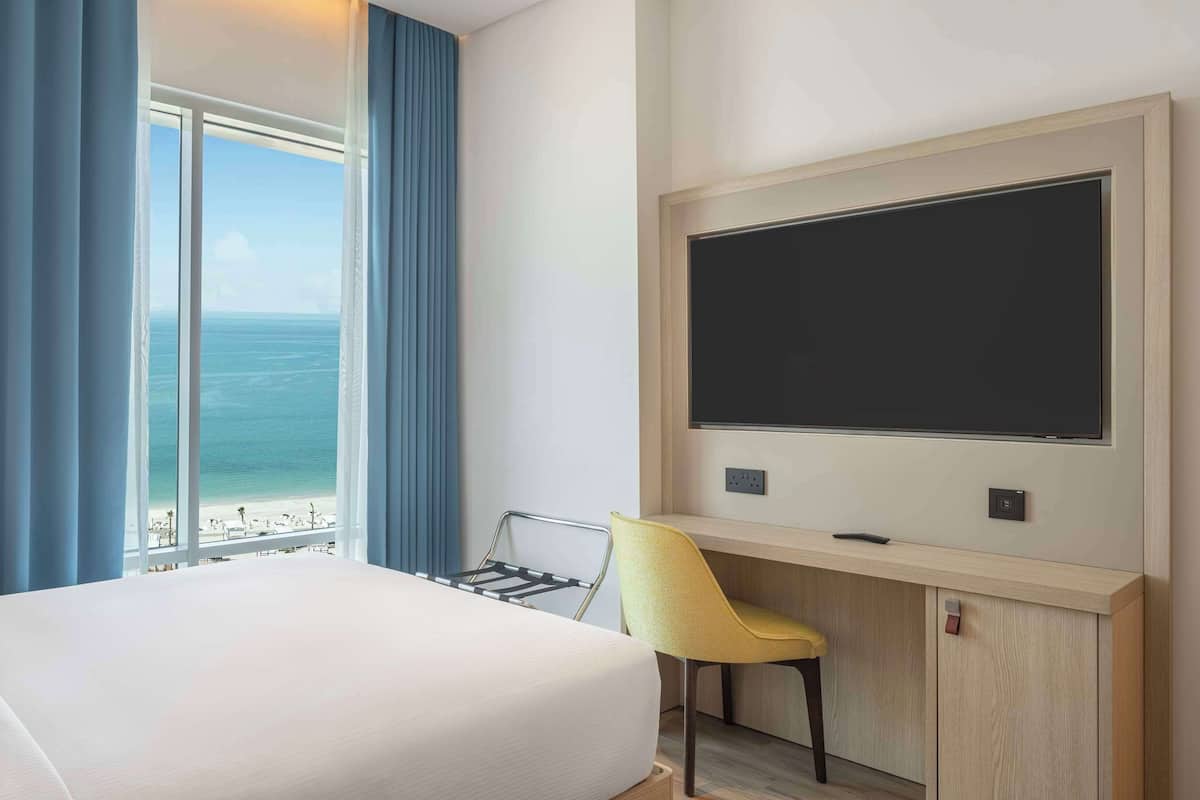 A hotel room with a bed, a desk with a chair, a large window with a view of the sea, and a flat-screen TV mounted on the wall.