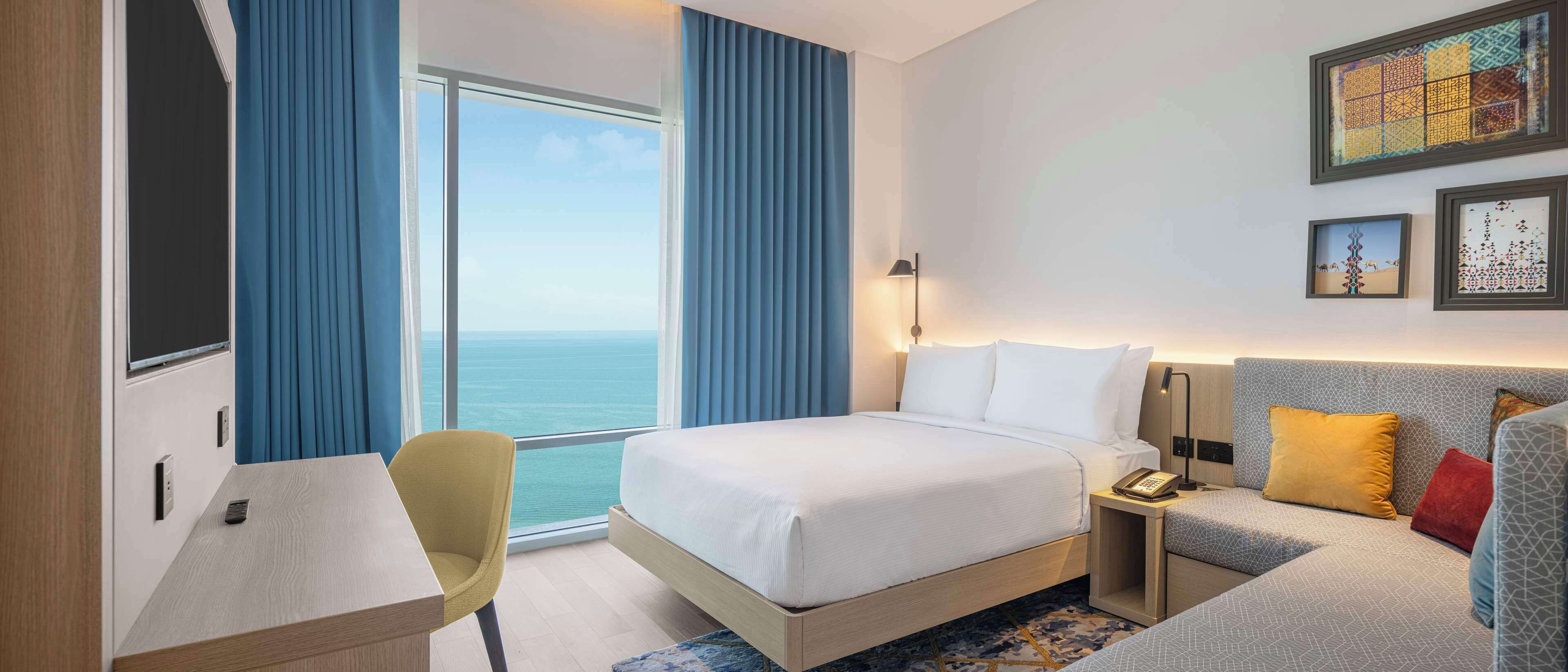 A hotel room with a large bed, a desk, a chair, a bedside table with a lamp, and a view of the ocean.