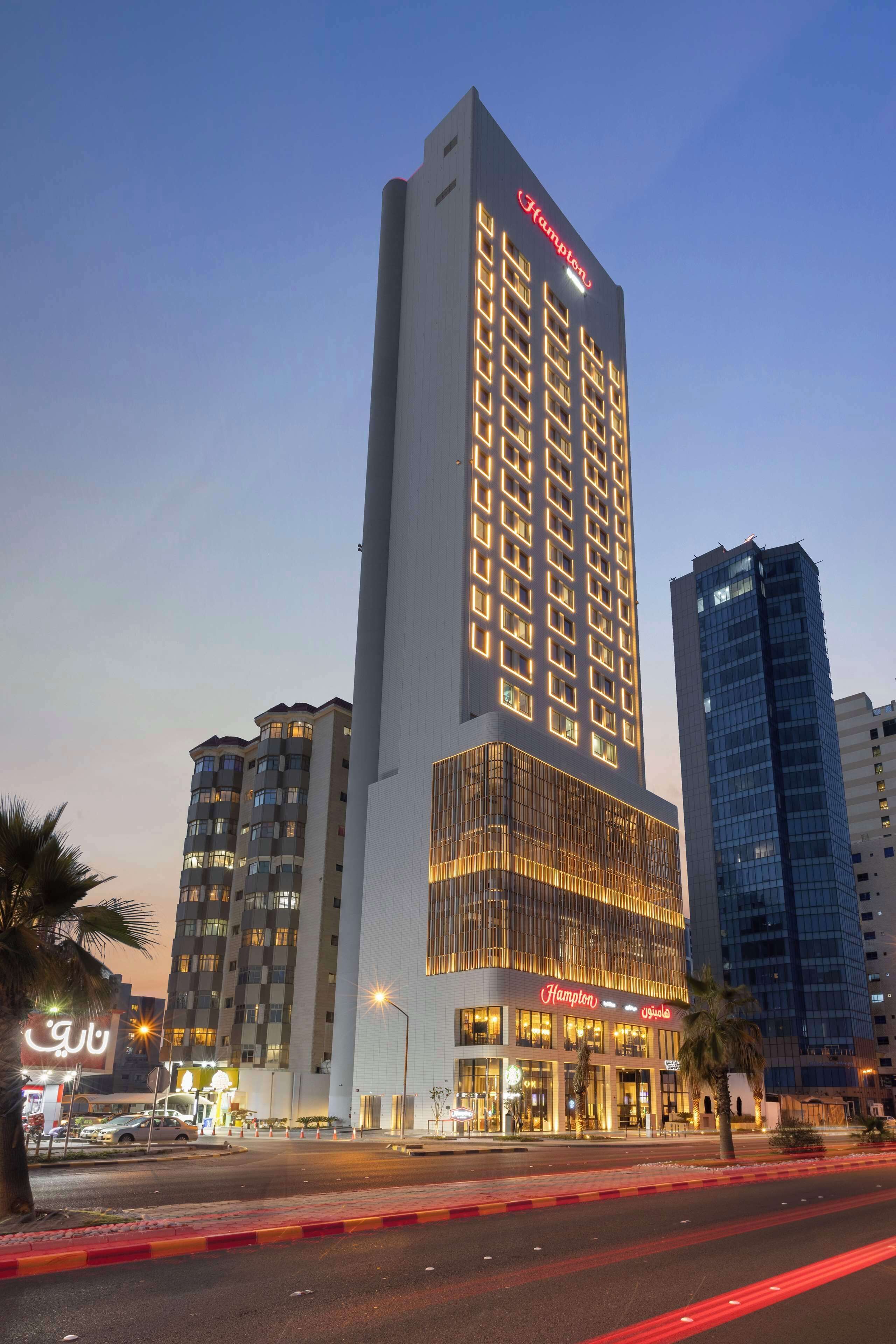 A tall hotel with a prominent 'Hampton' sign, featuring a modern glass facade and multiple floors.