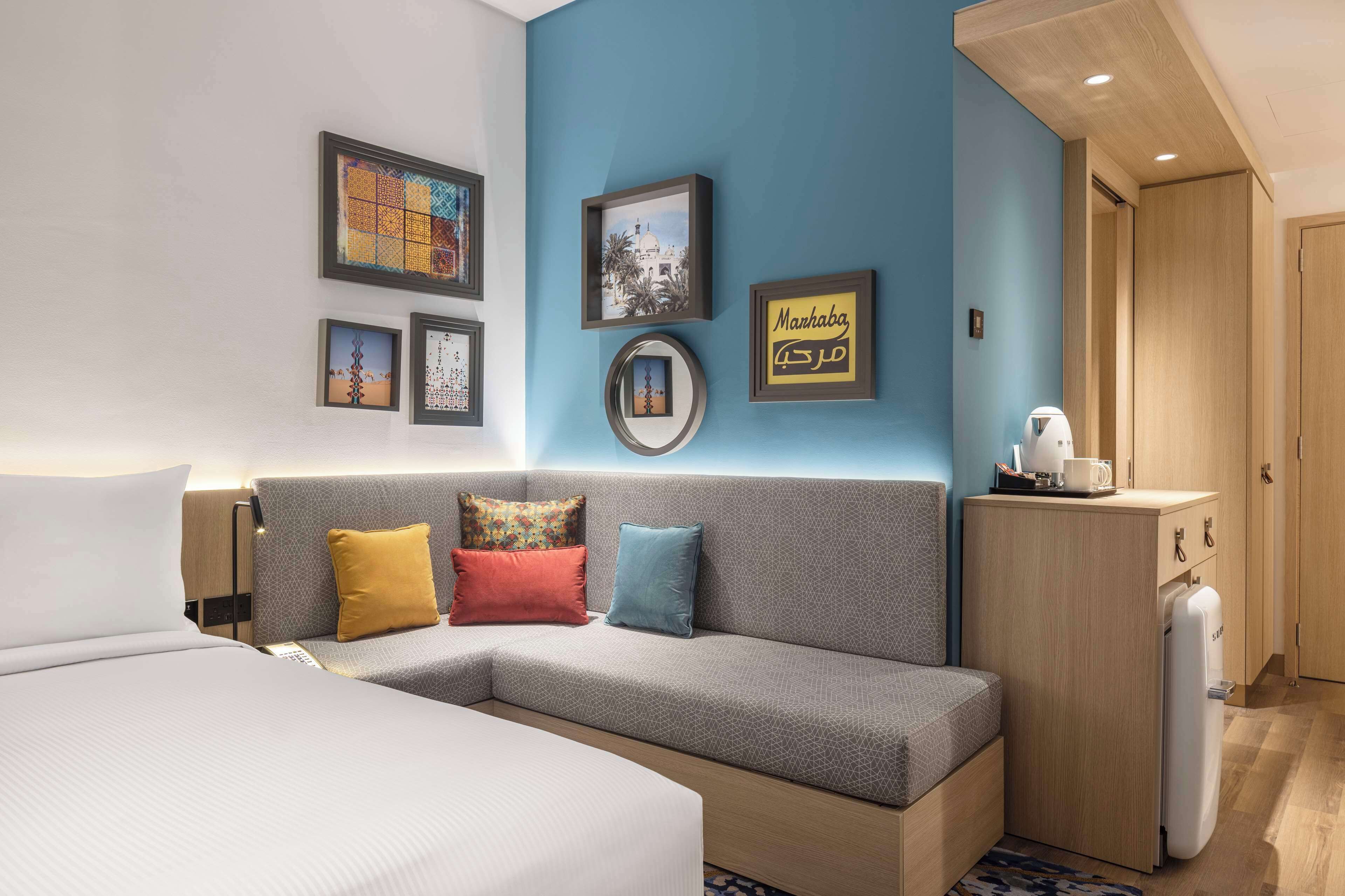 Una habitación de hotel moderna con un sofá gris, una cama, una pequeña cocina y obras de arte en las paredes.