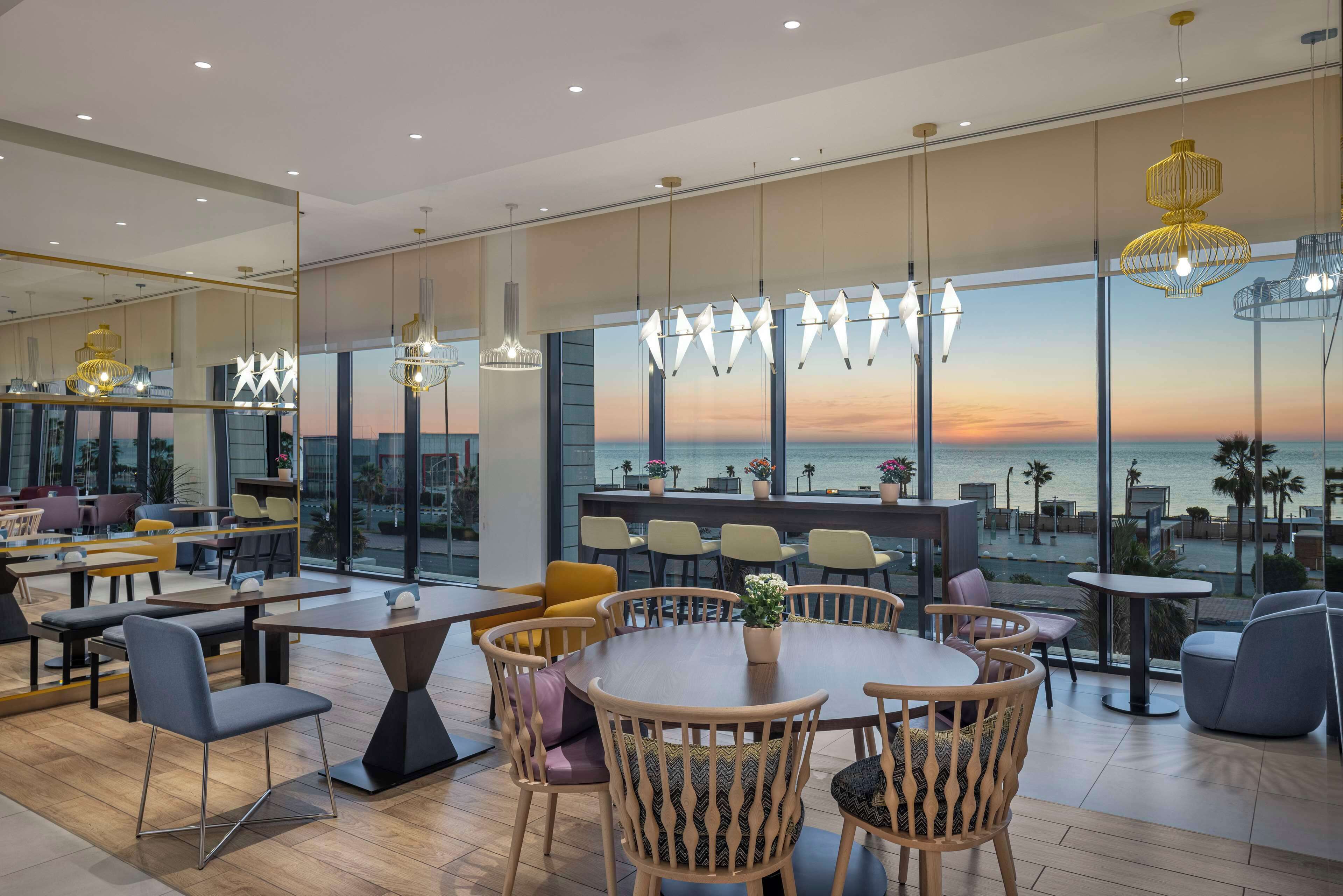 A modern dining area with round tables and chairs, large windows with a view of the sea, and hanging pendant lights.