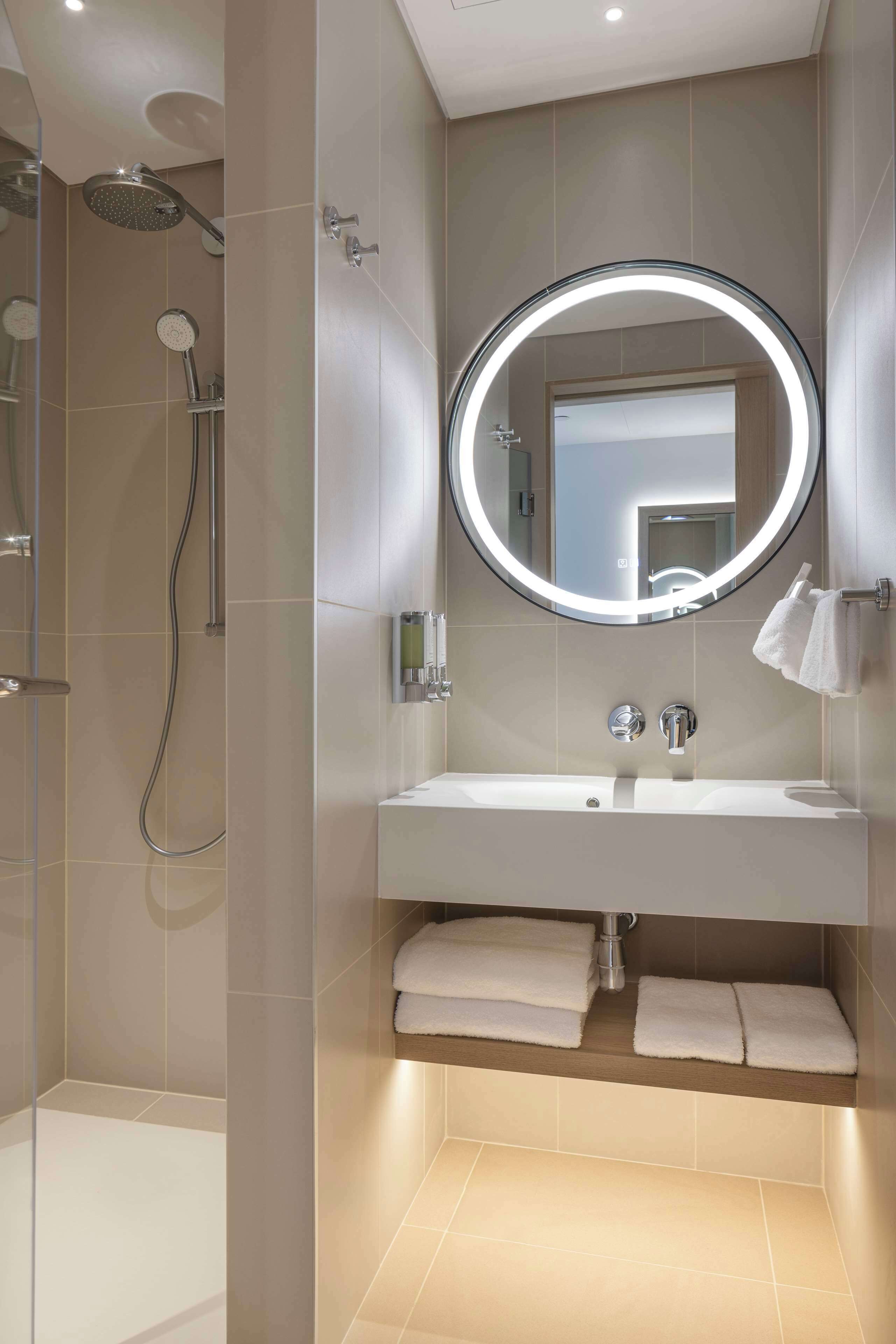 Un baño moderno con ducha a ras de suelo, un espejo grande con luz circular y un mueble de tocador con lavamanos y perchero.