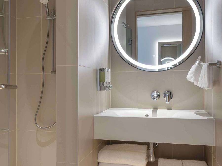 A modern bathroom with a walk-in shower, a large mirror with a circular light, and a vanity with a sink and towel rack.
