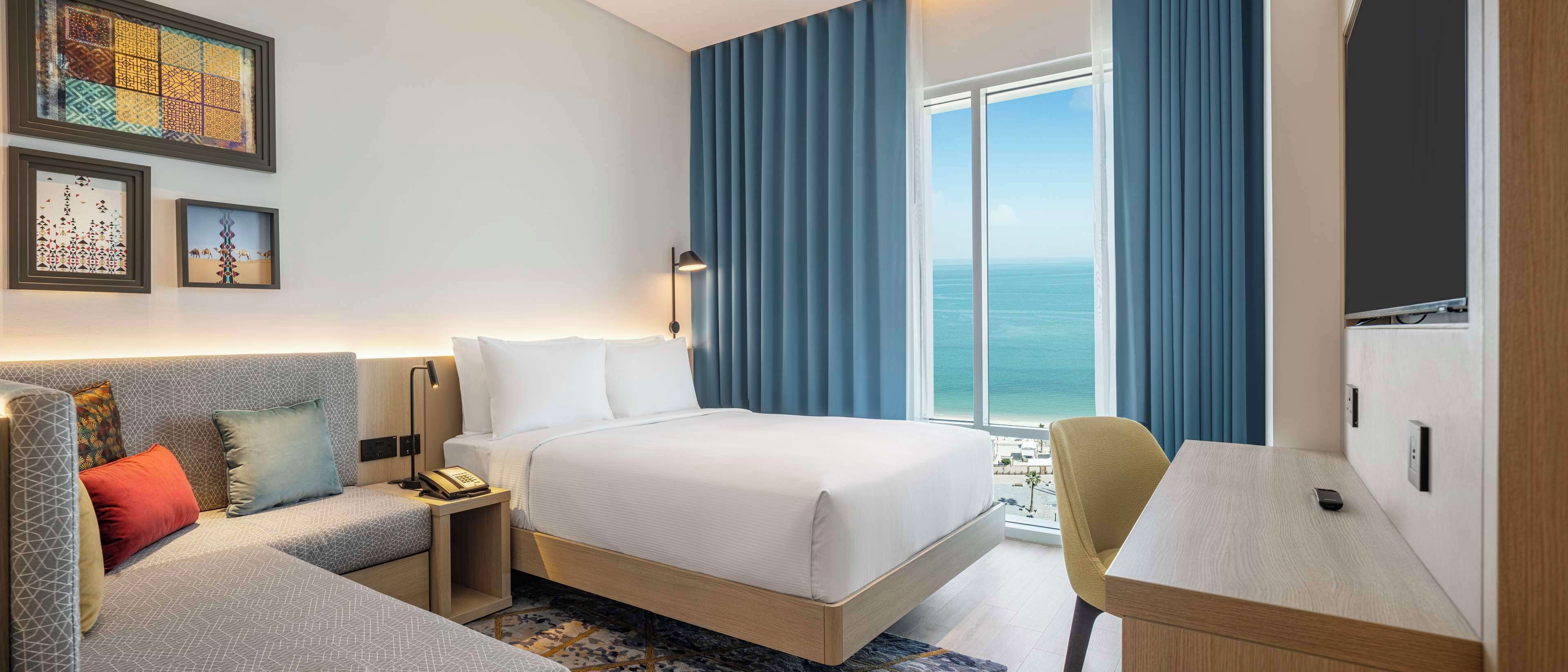 A hotel room with a bed, a desk, a sofa, and a view of the sea.