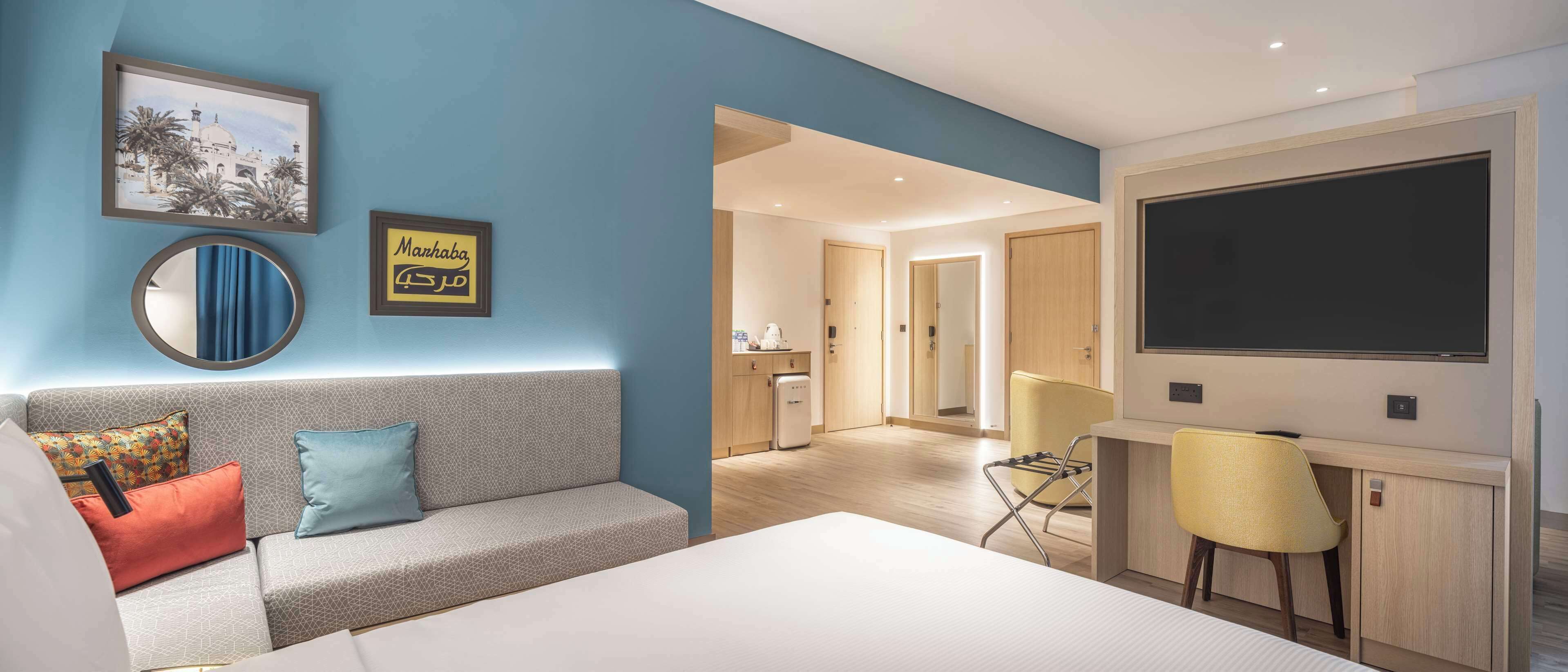 A modern hotel room with a sofa, a bed, a desk, and a TV. There are two doors leading to other rooms.