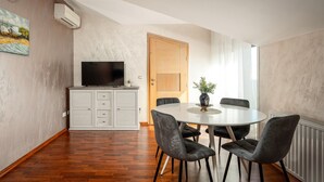 Uma sala de jantar com uma mesa redonda, quatro cadeiras, uma televisão sobre um móvel e uma porta que dá para outro cômodo.