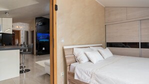 A modern bedroom with a wooden bed, a TV in the background, and a kitchen area visible through an open door. - Apartment Goran (Istria - Poreč) (Poreč)