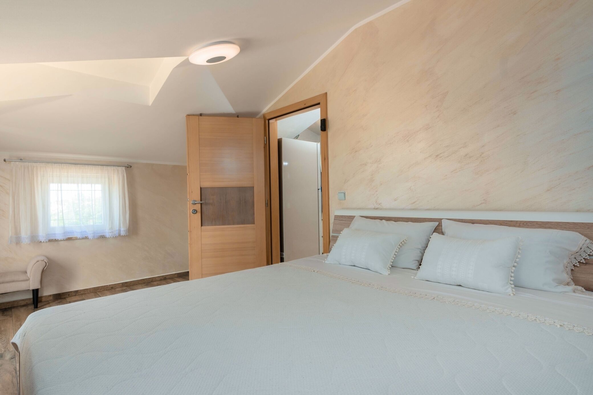 A bedroom with a large bed, a wooden wardrobe, a chair, and a window with curtains.