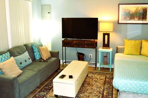 Una sala de estar con sofá, reposapiés, mueble para televisor y cama con colcha estampada.