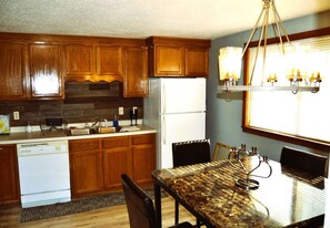 Una cocina con armarios de madera, lavavajillas empotrado, refrigerador, mesa de comedor con sillas y una araña.