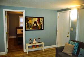 A living room with a blue wall, a white door, and a wooden floor. There is a white console table with decorative vases and a framed abstract painting above it.