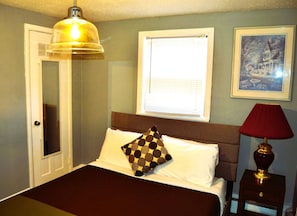 Un dormitorio con cama, lámpara de noche, ventana con persianas, puerta y un cuadro enmarcado en la pared.
